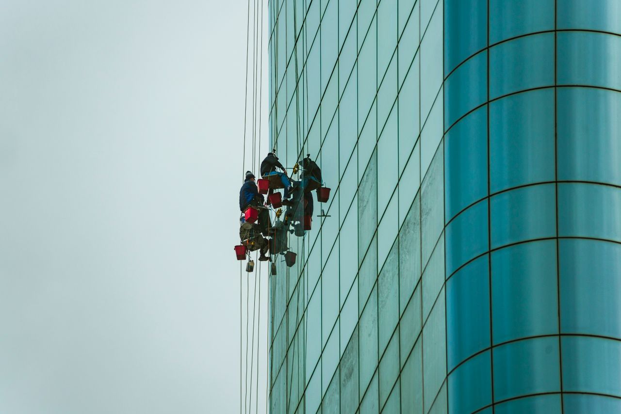 Window cleaners suspended on ropes working on a tall glass building facade.